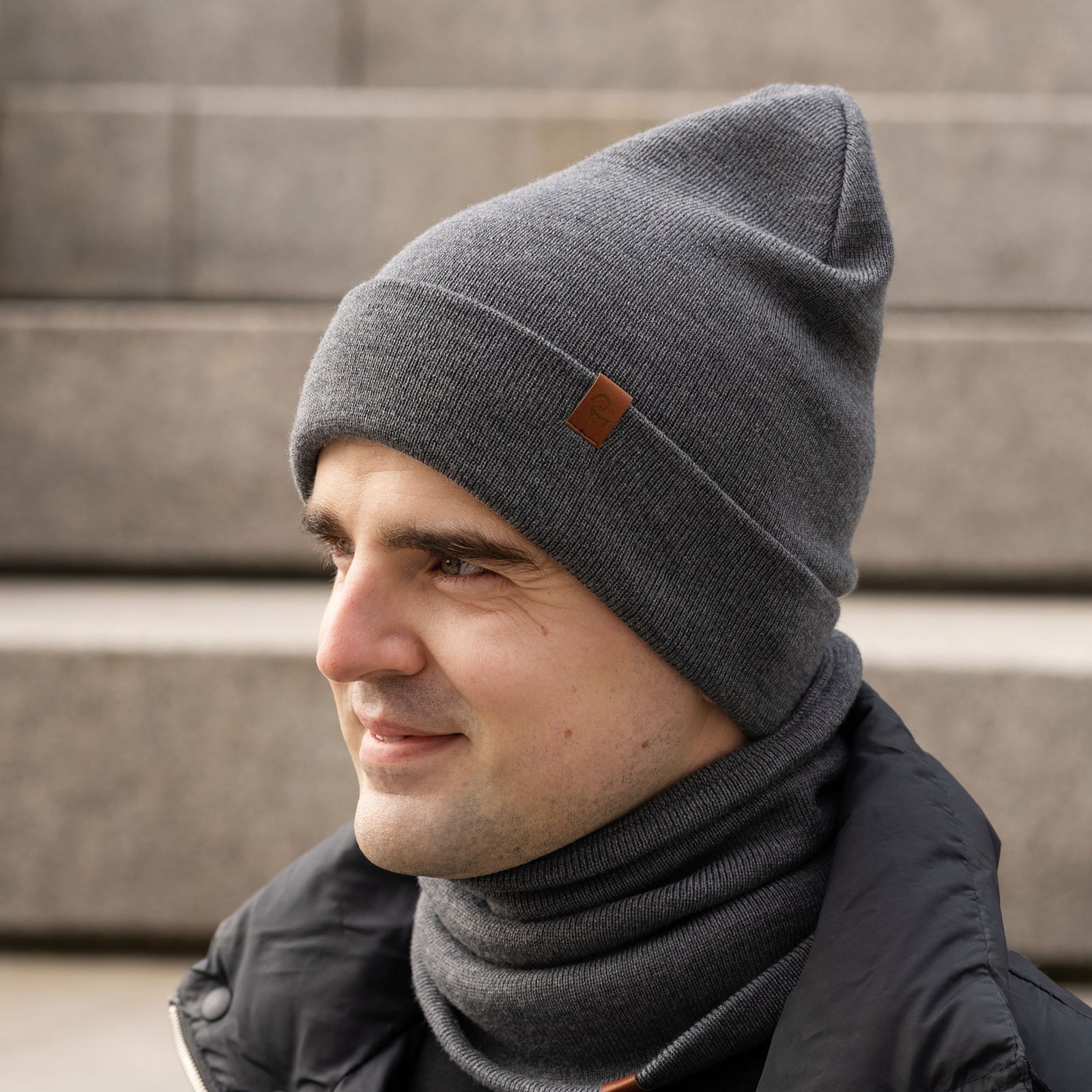 A person with light skin and dark hair is shown in profile, smiling slightly. They are wearing a black knit beanie with a small brown tag and a matching black knit neck gaiter, also with a small brown tag. They are wearing a black long-sleeved shirt. The background appears to be concrete steps or a similar textured surface in an outdoor setting.