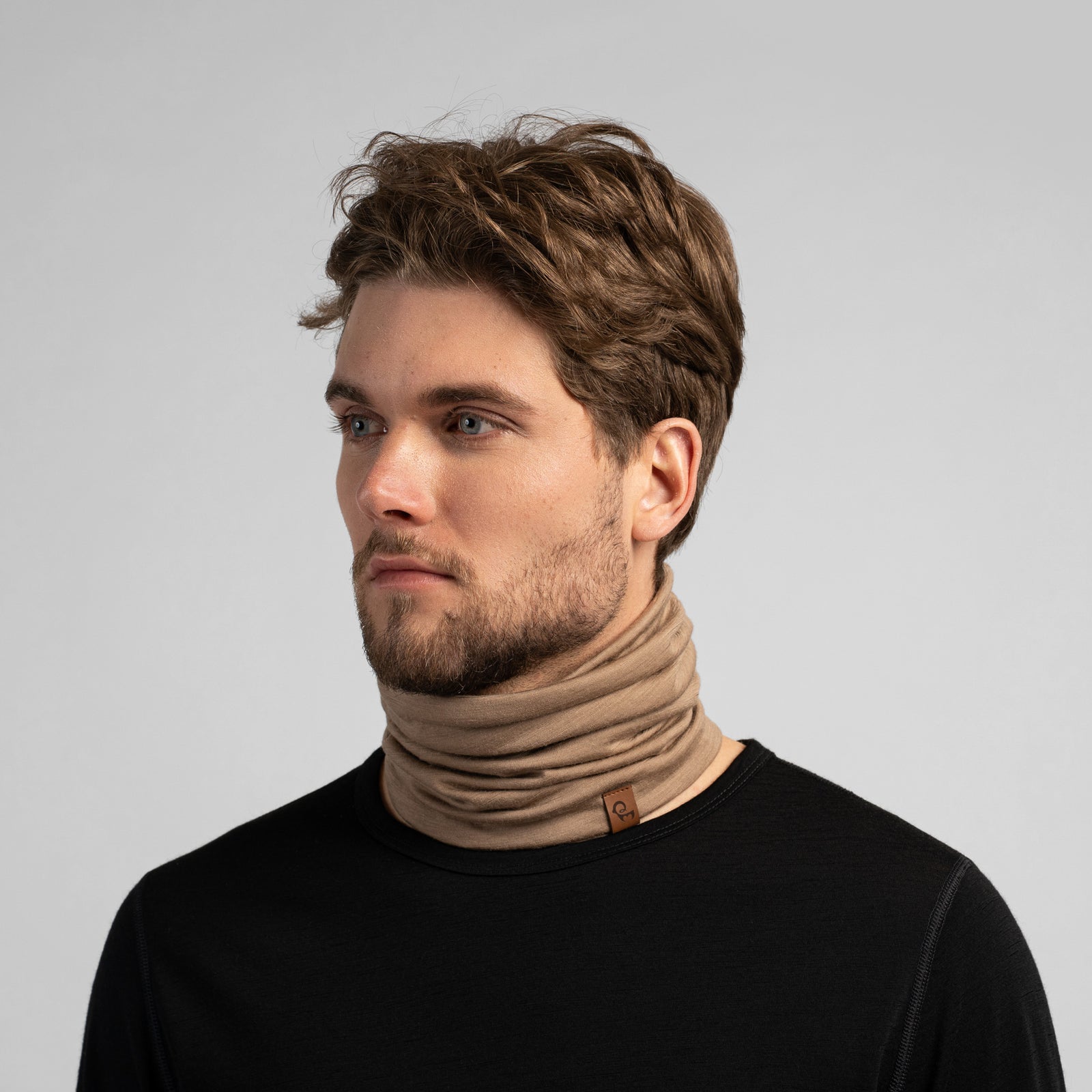A studio portrait of a man looking thoughtfully off to the left side of the frame. He is wearing a light beige neck gaiter pulled up over his nose, covering his lower face. The gaiter has a small brown leather logo patch on the bottom edge. He is wearing a solid black long-sleeved top against a plain light grey background.