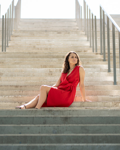 A woman with brown hair, wearing a bright red, sleeveless wrap dress, poses on indoor concrete stairs with metal railings.
