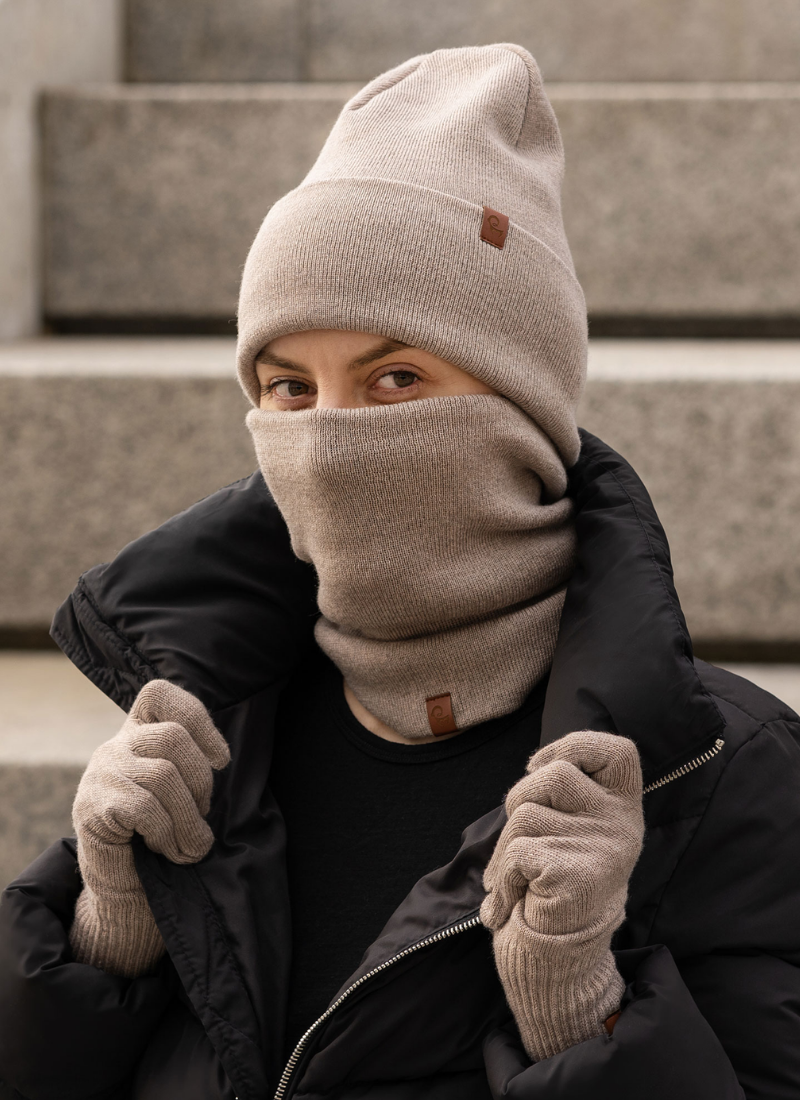 Person outdoors wearing a beige beanie, neck gaiter, and gloves with a black jacket.