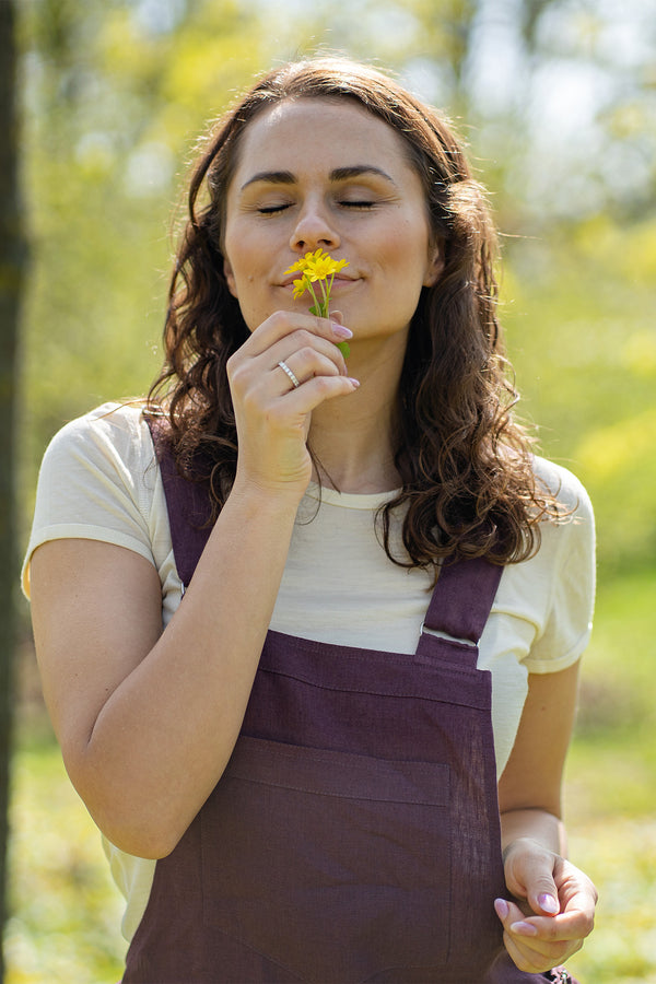 A woman with brown hair, wearing dark purple overalls over a light-colored t-shirt, stands outdoors between two tree trunks on grassy ground.
