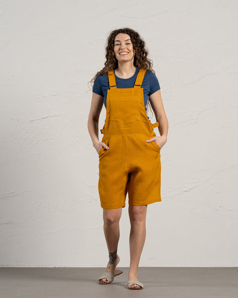 A woman wearing a blue t-shirt, mustard-colored overalls shorts, and light-colored sandals, posing against a textured off-white wall.