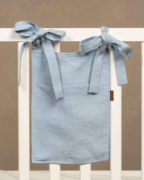 A light dusty blue linen baby bed organizer hanging from the top rail of a white wooden crib. It is secured by two wide fabric ties tied into bows. The organizer has a large front pocket with a small brown logo tag on the right seam. The background is a solid, warm taupe color.