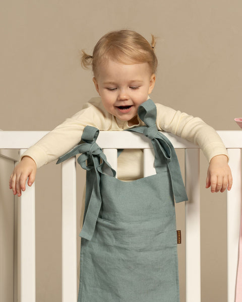 A smiling toddler leaning over the top rail of a white wooden crib. The child has blonde hair tied in tiny pigtails. Two linen baby bed organizers are hanging on the crib rail; a dusty sage green one in front of the child, and a pink one partially visible to the right. Both are tied to the rail with fabric bows. The background is a solid, warm taupe color.