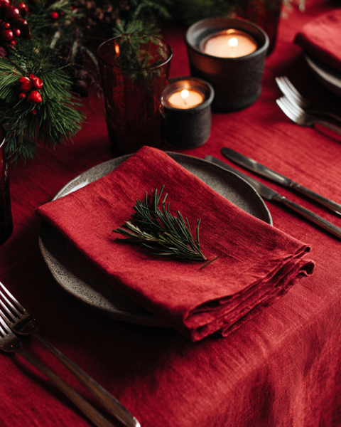 Festive Christmas table setting with a red linen napkin folded on a stone plate, decorated with a pine sprig and surrounded by candles and greenery.