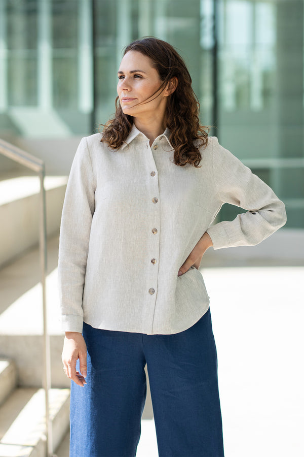 A woman with brown hair, wearing a natural linen button-front shirt and wide-leg white trousers, stands outdoors against a modern concrete structure.

