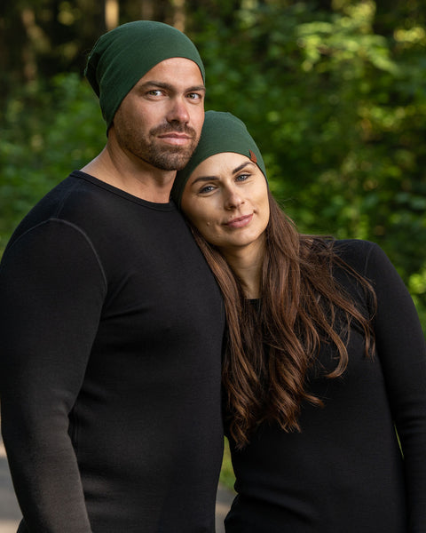 A half body upper body picture of a couple wearing unisex dark green menique merino headbands. They are smiling, the woman is leaning on the man&#39;s shoulder. They are also wearing black long sleeve merino tops.