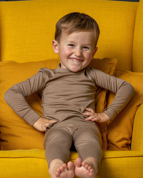 A boy smiling wide with his hands on his hips. He is wearing the menique merino long sleeve two piece set in beige color. he is sitting on a spicy yellow color ottoman.