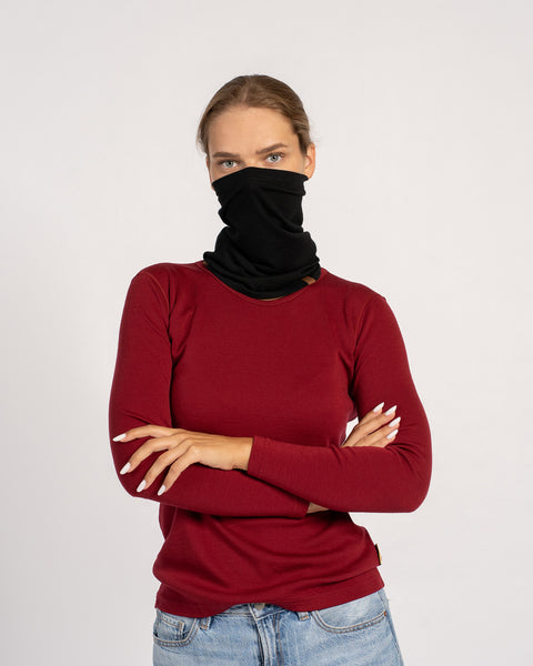 Woman wearing a black neck gaiter pulled up over her nose, dressed in a red long-sleeve shirt with crossed arms.