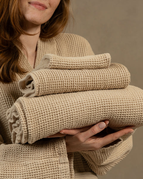 A close-up shot of a smiling woman's torso and hands as she holds a neatly stacked pile of three folded towels in varying sizes (a washcloth, hand towel, and bath towel). Both the towels and the robe she is wearing are made of a light beige, waffle-weave textured fabric. The background is a solid, warm taupe wall.