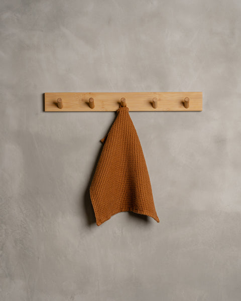 A gray wall with a wooden hanger. In the middle of it hangs a waffle face washcloth in almond brown color.