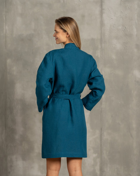 The menique women&#39;s linen bathrobe in cobalt blue color. The woman is standing in front of a gray wall and showing the bathrobe from the back. The adjustable tie is visible from the back.