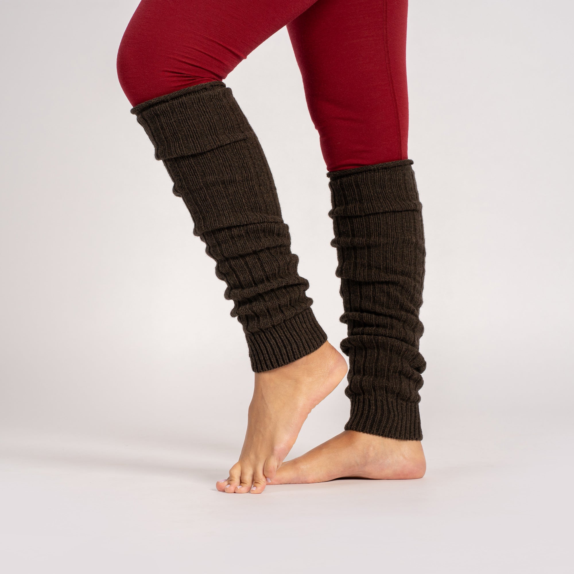 A woman sits on the floor in a studio setting, wearing an oversized beige mock-neck sweater and dark brown toeless leg warmers, looking at the camera.