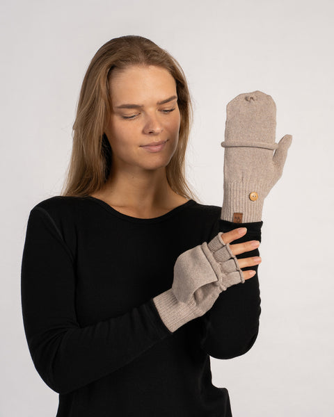 Frontal shot of the model looking down while holding up a creamy-beige convertible mitten (flap folded over) with the matching fingerless glove underneath.
