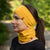 A person with long, dark hair pulled back in a ponytail is shown in a profile view, wearing a spicy yellow merino wool headband and a matching neck gaiter that covers their mouth and nose. They are wearing a black long-sleeved shirt, and the blurred background suggests an outdoor setting with green foliage.