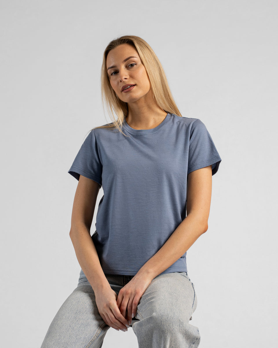 A studio portrait of a blonde woman sitting and leaning slightly forward, with her hands resting on her lap. She is wearing a regular fit, dusty blue short-sleeved crew-neck t-shirt and light-wash blue jeans. The background is a solid light grey.