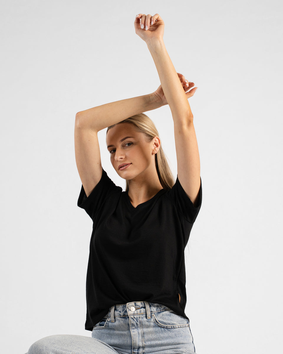 A studio portrait of a blonde woman looking directly at the camera. She is wearing a black short-sleeved V-neck t-shirt and light wash denim jeans, with her arms gracefully raised and hands resting together above her head against a plain light grey background.