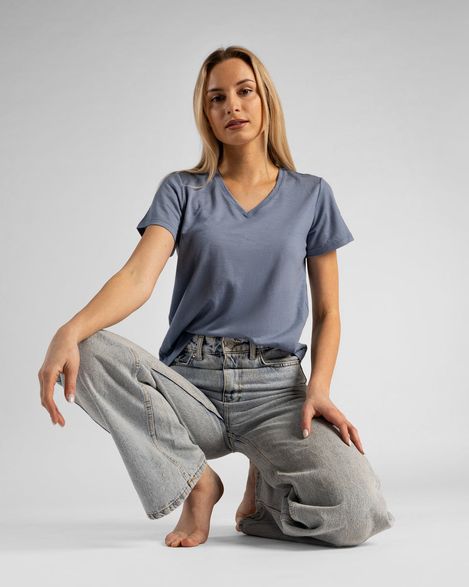 A studio shot of a barefoot blonde woman in a low crouch or kneeling pose on the floor, looking directly at the camera. She is wearing a dusty blue short-sleeved V-neck t-shirt and wide-leg light wash denim jeans, with her hands resting lightly on her knees against a plain light grey background.