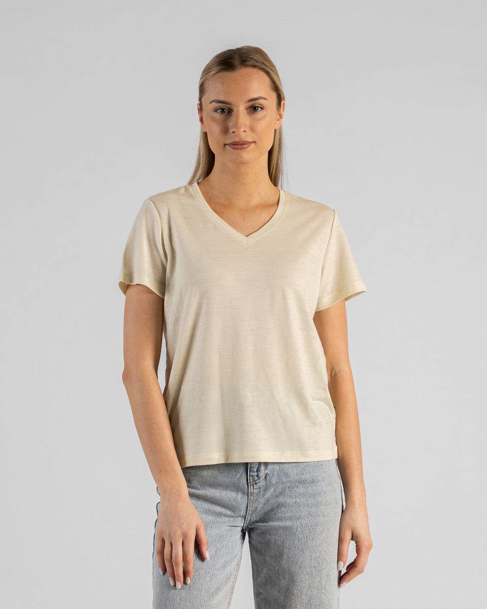 A studio portrait of a blonde woman standing and looking directly at the camera with her arms relaxed by her sides. She is wearing a regular fit, light cream natural-colored short-sleeved v-neck t-shirt and light-wash blue jeans against a plain light grey background.