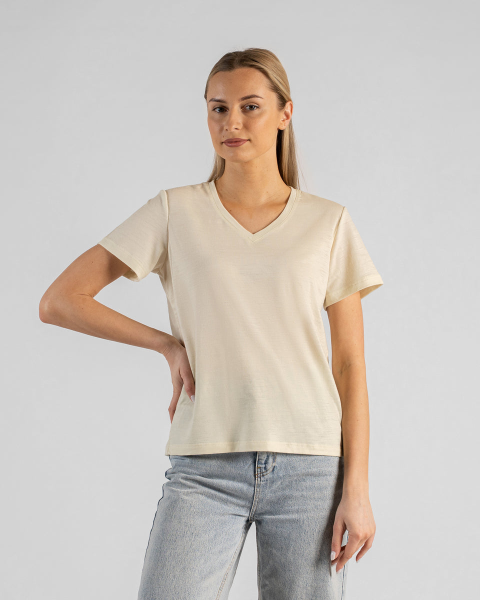 A studio portrait of a blonde woman standing with one hand resting on her hip, looking directly at the camera with a subtle smile. She is wearing a natural off-white short-sleeved V-neck t-shirt and light wash denim jeans against a plain light grey background.