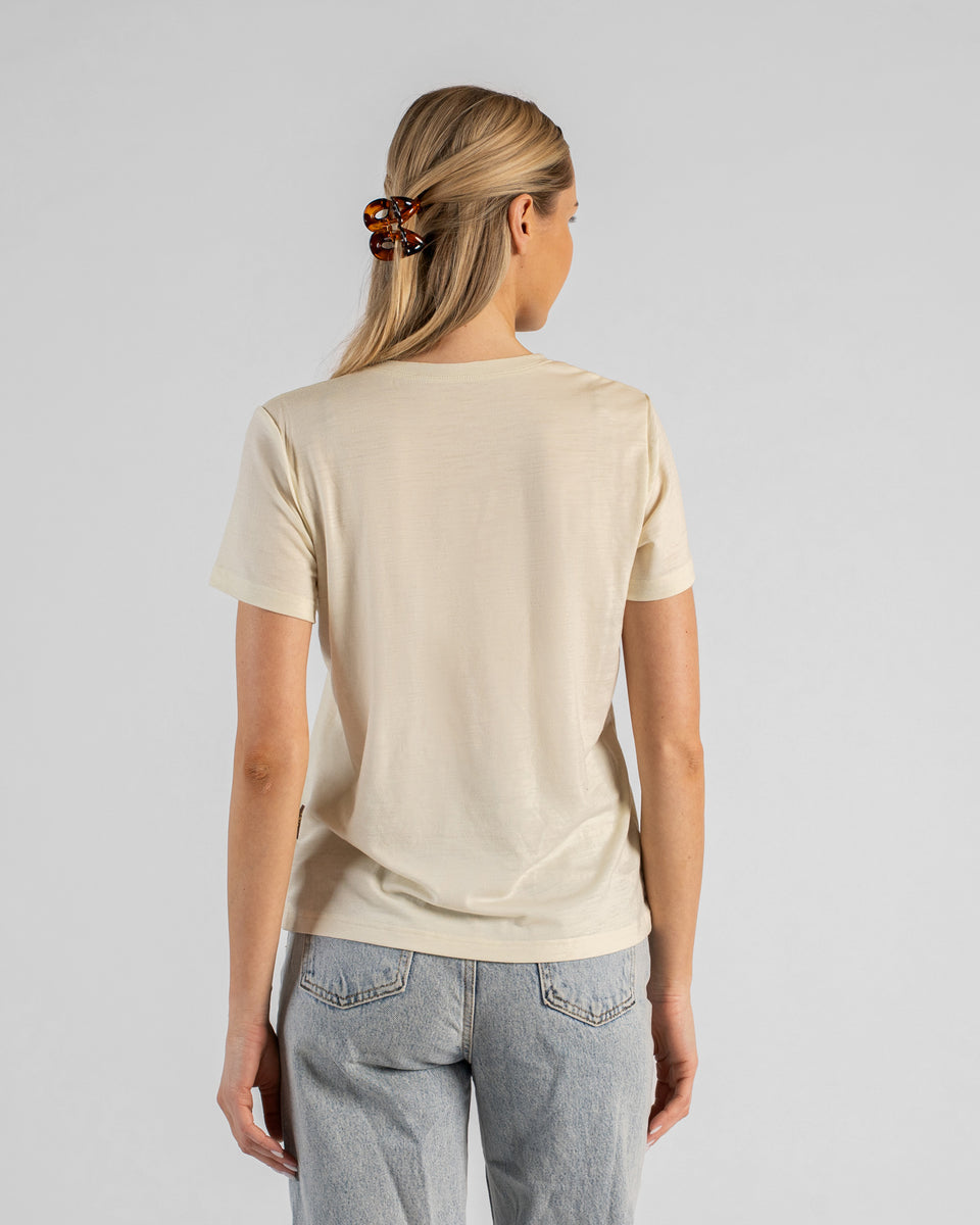 A back view studio shot of a blonde woman standing against a plain light grey background. She is wearing a natural off-white short-sleeved V-neck t-shirt and light wash denim jeans, with her hair styled half-up using a brown tortoiseshell claw clip.
