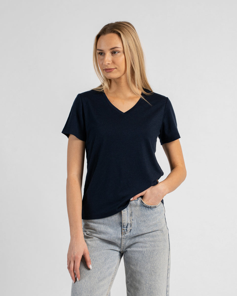 A studio portrait of a blonde woman standing and looking slightly off to the left against a plain light grey background. She is wearing a dark navy blue short-sleeved V-neck t-shirt and light wash denim jeans, with one hand resting in her front pocket.