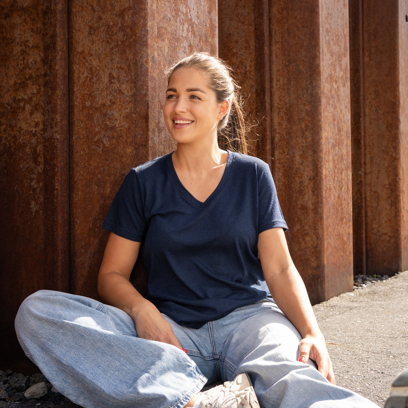 A smiling woman leaning against a rust-colored corrugated metal wall, wearing a dark blue v-neck t-shirt and blue jeans.