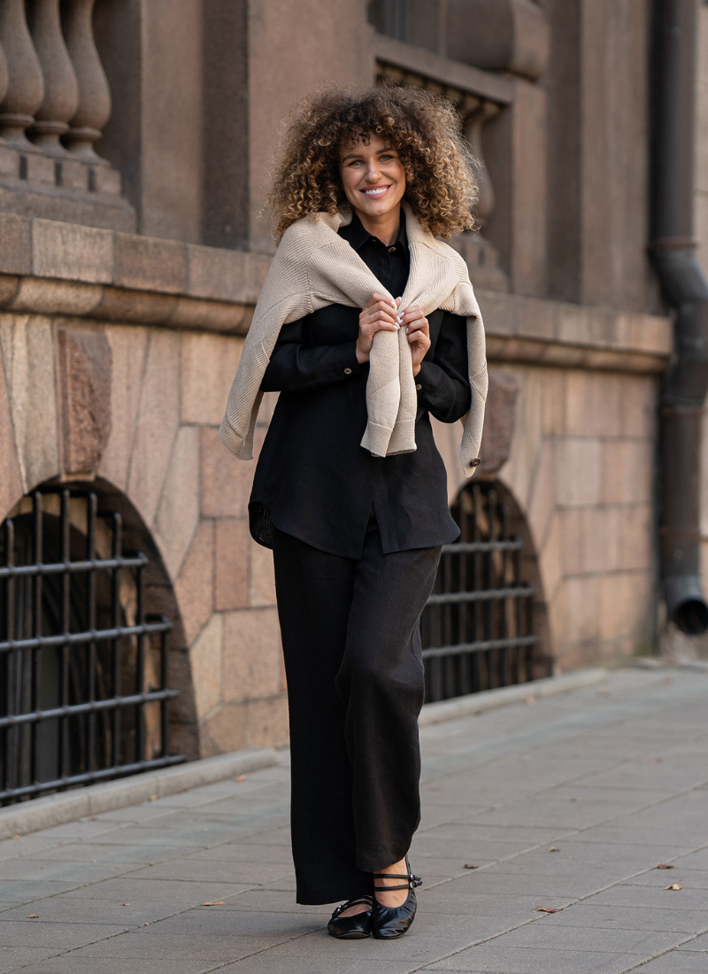 Woman wearing a black linen outfit with a light linen sweater draped over her shoulders, walking along a stone city street, showcasing relaxed elegance and breathable natural fabric.