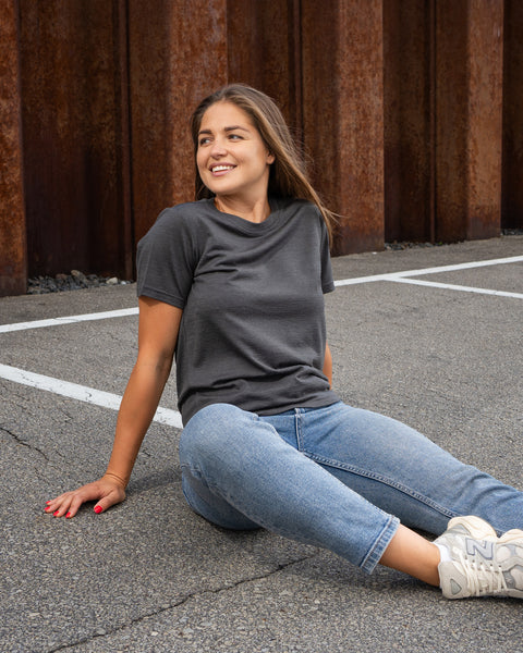 A smiling woman sitting on a paved surface, leaning back on her right arm and looking off to the side. She is wearing a charcoal grey crew neck t-shirt, blue jeans, and light grey sneakers, with a rusty corrugated metal wall in the background.