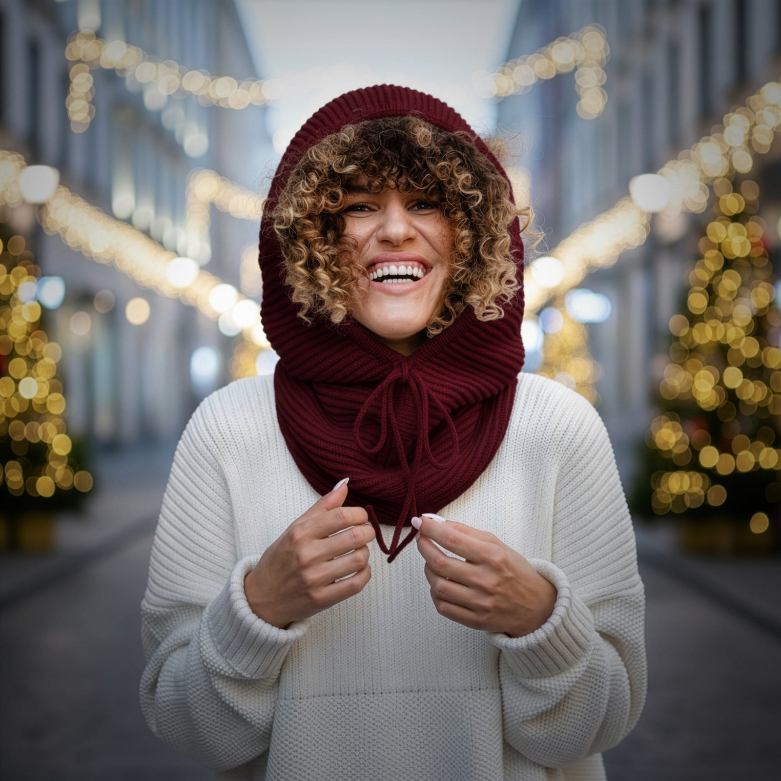 Person wearing a red hood and scarf in a festive street setting with blurred lights and Christmas trees.