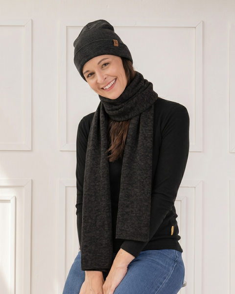 A smiling woman sitting and looking directly at the camera. She is wearing a black knit beanie with a brown leather logo patch and a matching long black knit scarf wrapped around her neck. She is dressed in a black long-sleeved top and blue jeans, posed against a white paneled wall.