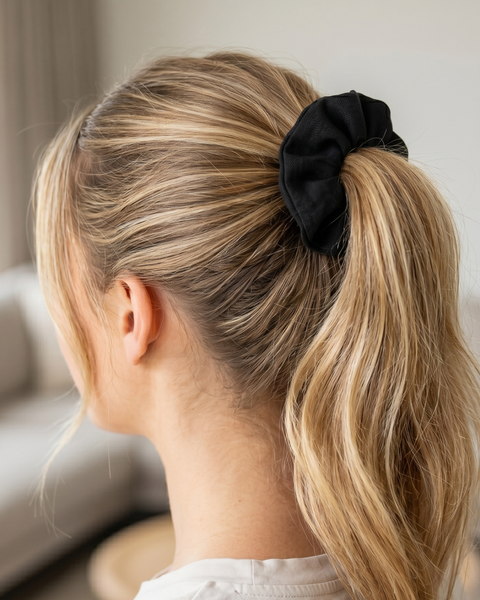 The back of a woman's head, showing her blonde hair pulled back into a mid-height ponytail secured with a black fabric scrunchie. The background is a softly blurred modern living room with a beige sofa.