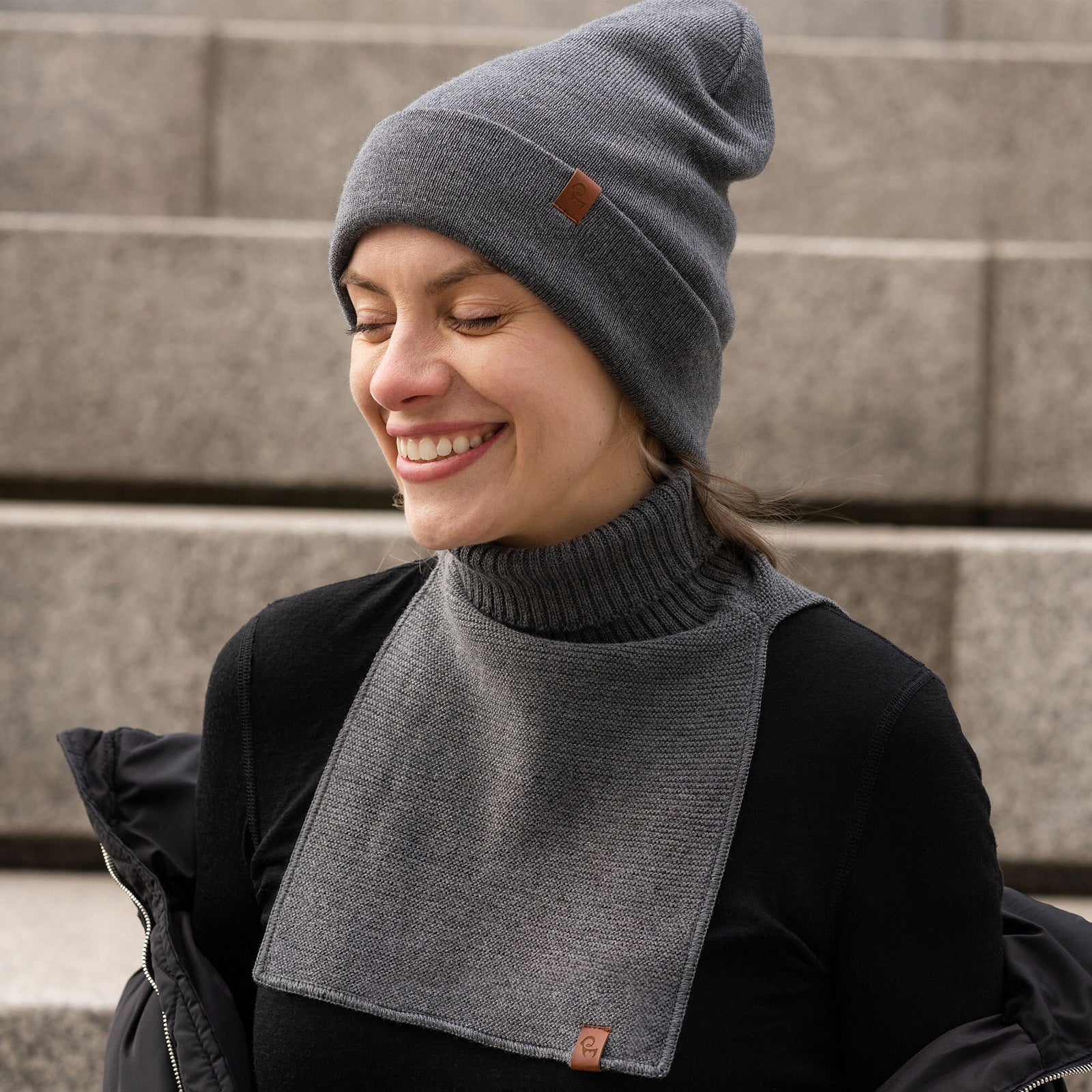 A woman with a fair complexion is pictured outdoors, smiling and looking off-camera to her right. She's wearing a light brown or taupe ribbed knit beanie with a small, square brown logo patch on the left side of the cuff. Around her neck, she has a matching knit neck warmer; this piece features a thick ribbed turtleneck and a rectangular, flat-knit bib that extends down her chest. This neck warmer also has a small brown logo patch at the bottom corner of the bib.
