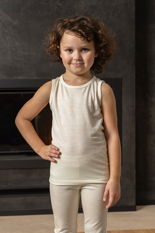 A young child with curly brown hair stands in front of a dark, textured wall with a modern fireplace opening. The child is wearing a natural sleeveless top and matching pants. They are looking directly at the camera with a slight smile, with one hand on their hip.