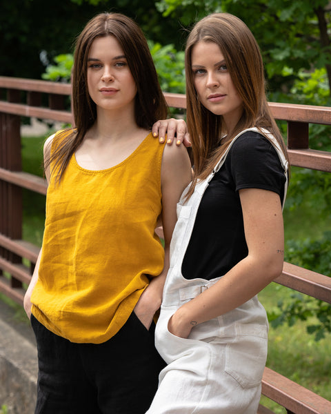 Two girls posing in a city street. One is wearing spicy yellow linen tank top Hana and pure black pants, other one is wearing merino wool top black color and pure white linen jumpsuit.