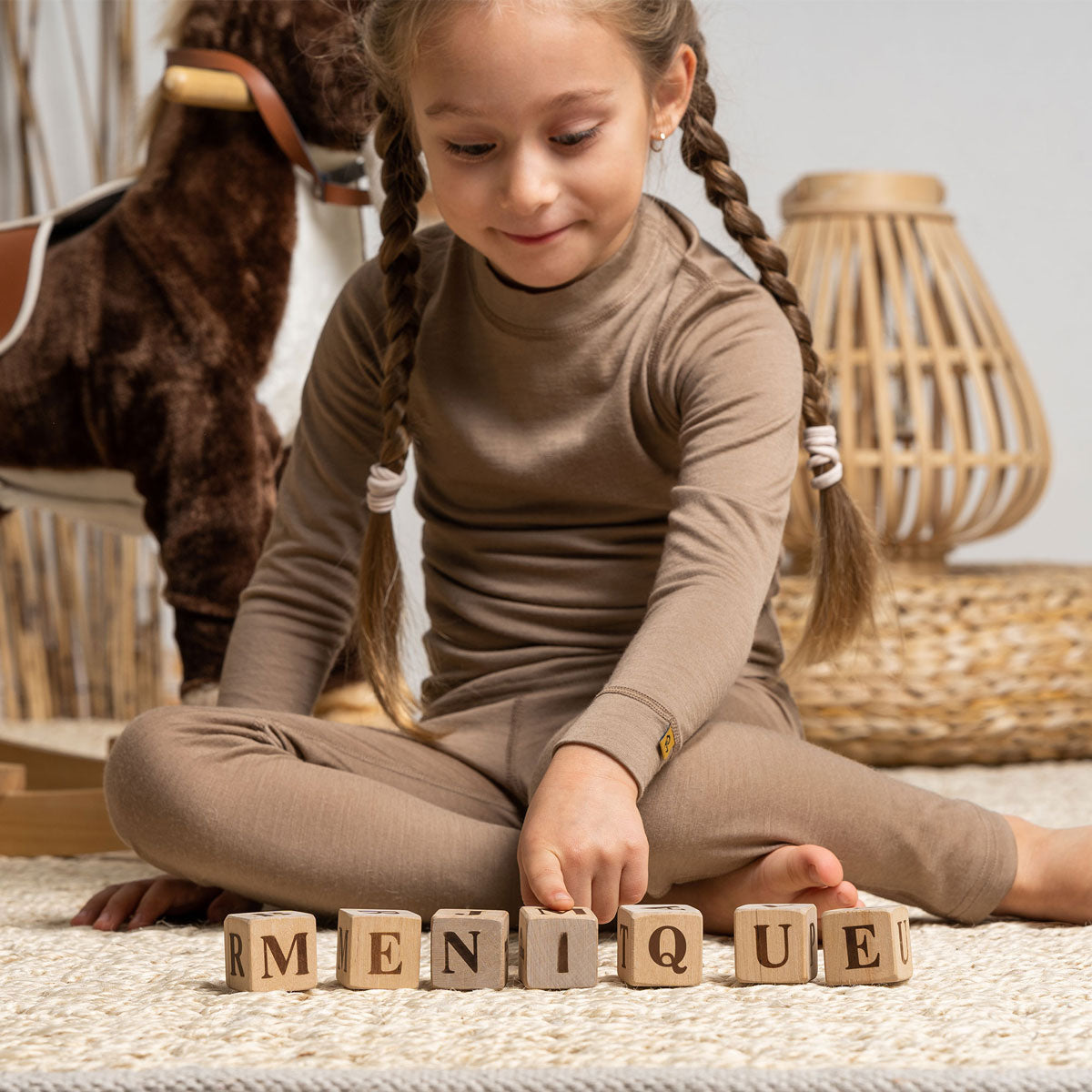 A girl, holding a teddy bear, wearing the menique Merino 160 Pants Beige color. She is standing on a bed, smiling. 